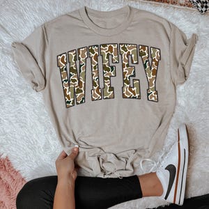 Pode incluir: Camiseta bege com a palavra "WIFEY" em um padrão de camuflagem na frente. A camiseta é combinada com leggings pretas e tênis brancos com um logotipo preto e detalhes marrons. A camiseta é dobrada na parte inferior.