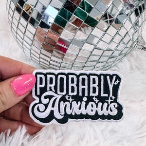 May include: A black and white embroidered patch with the text "Probably Anxious" in a white cursive font with glitter accents.