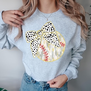 May include: A light gray sweatshirt featuring a baseball-themed design. The design includes a baseball with a gold glitter outline, a dalmatian print bow, and red stitching. The sweatshirt is a casual, everyday garment.