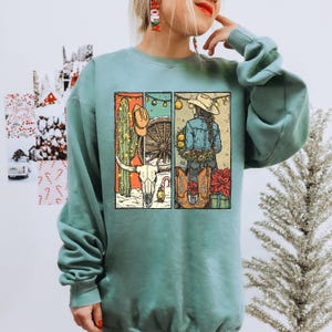 Puede incluir: Sudadera turquesa con un diseño gráfico de temática vaquera. El diseño incluye un cactus, un sombrero de vaquero, una rueda de carro, una calavera de cuernos largos y una vaquera con botas y flores. El diseño es en tonos naranja, verde y marrón.