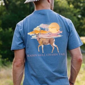 May include: Blue t-shirt with a deer graphic, sunset, and text "RAISIN HELL CHASIN TAIL." A camouflage baseball cap is worn. The design features a deer, sun, and birds in flight.