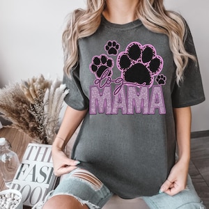 May include: Dark gray t-shirt with a graphic design featuring black paw prints outlined in pink and the words "dog MAMA" in a glittery pink font. The shirt is a casual style.
