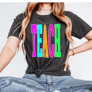 May include: A black t-shirt with the word "TEACH" printed in rainbow colors. The letters are outlined in white.