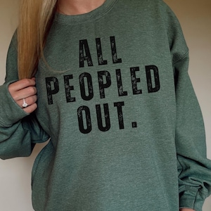 May include: Green sweatshirt with the text "ALL PEOPLED OUT" in black, distressed lettering. The sweatshirt is a solid color and has a crew neck. The sleeves are long and the material appears to be a soft fabric.