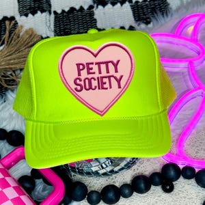 May include: A neon green trucker hat with a pink heart-shaped patch. The patch features the words "Petty Society" in a dark pink font. The hat has a mesh back and a curved brim.