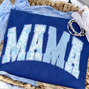 May include: A navy blue t-shirt with the word "MAMA" in light blue, floral, and polka dot patterned letters. The letters have a scalloped edge. A pearl bracelet and a light blue bow are in the background.