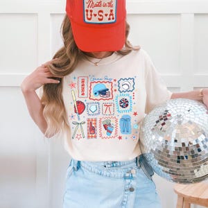 May include: Cream-colored t-shirt with baseball-themed graphics, including a baseball bat, helmet, and glove. The shirt also features the text "Game Day" and "Baseball". A red baseball cap with "Made in the U.S.A." is worn.