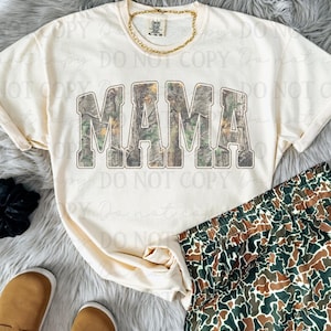 May include: Cream-colored t-shirt with the word "MAMA" in a camouflage design. The shirt is paired with camouflage shorts and a gold chain necklace. Tan loafers and a black scrunchie are also visible.