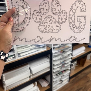 May include: A white sheet with the words "DOG MAMA" spelled out in black rhinestones. The letter "D" is stylized with a leopard print pattern, as is the paw print in the center. The letter "G" is also stylized with a leopard print pattern.