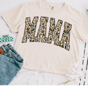 May include: A beige t-shirt with a camo print "MAMA" graphic.
