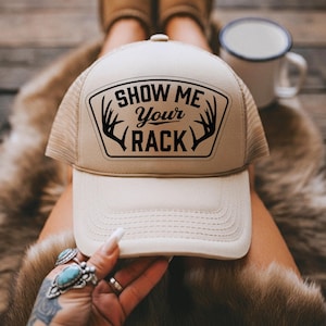 May include: Tan trucker hat with a white front panel and mesh back. The hat features the text "SHOW ME Your RACK" in black, framed by antler illustrations. A casual accessory.