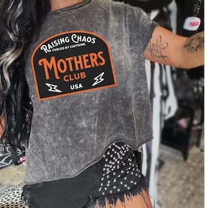 May include: A gray, distressed t-shirt with a black and orange patch that reads "Raising Chaos Fueled by Caffeine Mothers Club USA".
