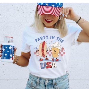 Könnte beinhalten: Weißes T-Shirt mit einem rot-weiß-blauen amerikanischen Flaggenmuster. Das Shirt zeigt einen Cartoon-Hotdog und ein Bierglas, die sich die Hände halten, mit dem Text "Party in the USA".