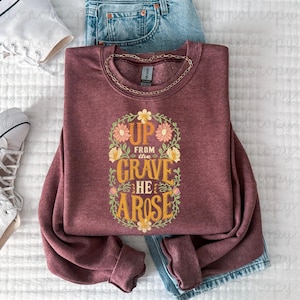 May include: Burgundy crewneck sweatshirt with the text "UP FROM THE GRAVE HE AROSE" in a floral wreath design. The sweatshirt is folded on a white surface with a pair of blue jeans and white sneakers.