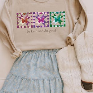 May include: A tan sweatshirt with a colorful butterfly quilt design and the text "be kind and do good". The sweatshirt is paired with a light blue floral skirt and white cowboy boots.