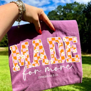 May include: A purple t-shirt with the words "MADE for more" in a retro font. The letters are filled with an orange and pink checkerboard pattern and small white flowers. The text "Ephesians 2:10" is below.