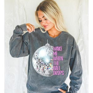 May include: A gray sweatshirt with a large silver disco ball graphic and the text "Wake Me When The Ball Drops" in pink.