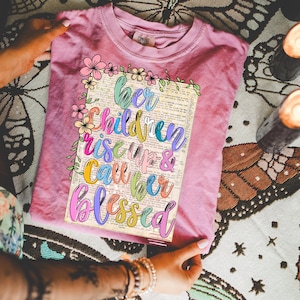 May include: A dusty rose-colored t-shirt with a crew neck. The shirt features a rectangular graphic with the words "Her Children Rise Up & Call Her Blessed" in colorful script. Floral and leaf accents surround the graphic.