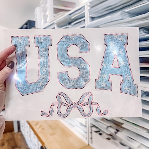 Puede incluir: Una hoja blanca con la palabra "USA" en pedrería azul, con contornos rojos y detalles de estrellas. Un diseño de lazo, también en pedrería roja y azul, se encuentra debajo de las letras.