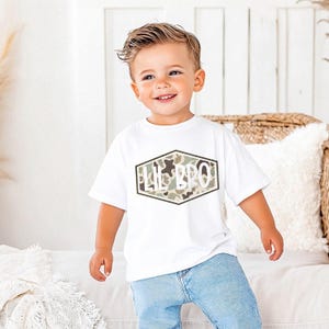 May include: A white t-shirt with the text "LIL BRO" in a camouflage design. The text is inside a hexagonal shape. The shirt is worn by a young child wearing blue jeans.