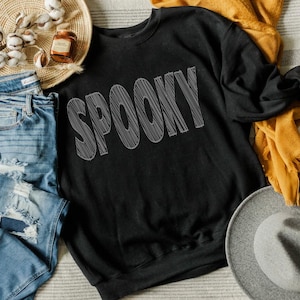 May include: Black crew neck sweatshirt with the word "SPOOKY" in gray, scribbled lettering. The sweatshirt is displayed with a straw hat, a yellow scarf, and a pair of blue jeans.