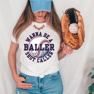 May include: White t-shirt with a blue and red baseball graphic and the text "Wanna Be A Baller Shot Caller".