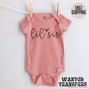 May include: A light pink baby onesie with the words "lil sis" in black script. The onesie is hanging from a clothesline with wooden clothespins. The onesie has a snap closure at the bottom. The brand name "Gerber" is visible.