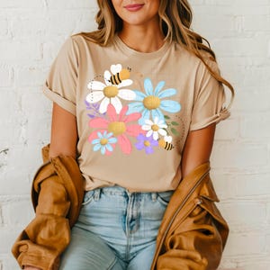 May include: Beige t-shirt featuring a colorful floral design with bees. The graphic includes daisies in white, pink, blue, and purple, with yellow centers. The shirt has short sleeves and a relaxed fit, suitable for casual wear.