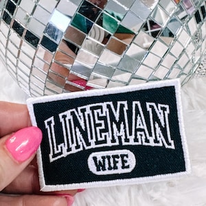 Peut inclure: Écusson rectangulaire noir avec texte brodé blanc "LINEMAN WIFE". Le patch a une bordure blanche. Il est présenté sur une surface blanche duveteuse, avec une boule à facettes en arrière-plan.