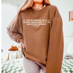 May include: A brown sweatshirt with the text "IF YOU'RE HANGING ON BY A THREAD, MAKE SURE IT'S THE HEM OF HIS GARMENT. MATTHEW 8:20-22" printed on the front.