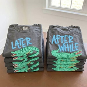 May include: Stacks of gray t-shirts with alligator graphics. One stack has the word "LATER" in blue, and the other says "AFTER WHILE" in blue. The alligators are green with red accents.