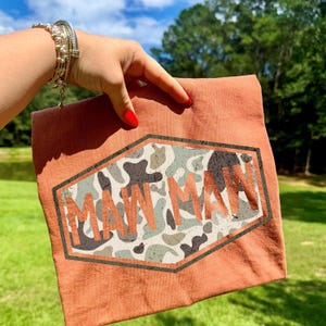 May include: A terracotta-colored t-shirt featuring a camouflage design within a hexagonal frame. The word "MAW MAW" is printed in a distressed, orange font across the camouflage pattern. The shirt is held up against a backdrop of green grass and a blue sky.