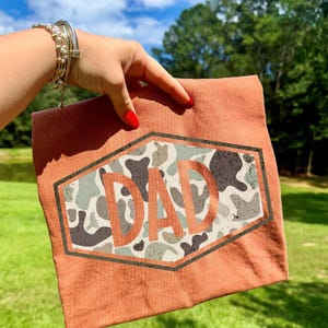 May include: A rust-colored t-shirt featuring a camouflage design within a hexagonal frame. The word "DAD" is prominently displayed in a bold, distressed font, making it a perfect gift for Father's Day or any special occasion.