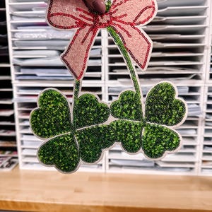 May include: A decorative sequined bow and shamrock applique. The bow is light pink with a red outline and center. The shamrocks are green with a white outline. The applique is held up by a hand.