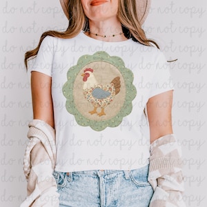 Puede incluir: Camiseta blanca con un diseño circular de un pollo. El pollo tiene una cresta roja, plumas marrones y un cuerpo con estampado floral. El diseño está enmarcado por un borde verde festoneado con pequeños detalles florales.