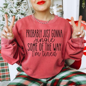 Puede incluir: Una sudadera de color rojo anaranjado con el texto "PROBABLY JUST GONNA jingle SOME OF THE WAY I'm tired" en negro. La sudadera forma parte de una colección con temática navideña. La persona lleva un collar y tiene pintalabios rojo.