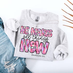 May include: A light gray sweatshirt with the text "HE MAKES all things NEW" in a colorful, watercolor-style design. The text "Revelation 21:5" is below the word "NEW". A silver necklace is on the sweatshirt.
