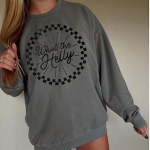 May include: A gray sweatshirt with the text "What the Helly" in a black, circular design. The design includes a checkered border and radiating lines. The sweatshirt has long sleeves and a crew neck.
