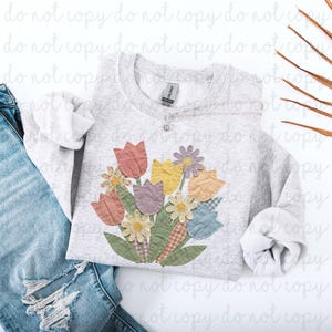May include: A light gray sweatshirt featuring a colorful floral applique design. The applique includes tulips, daisies, and leaves in shades of pink, yellow, purple, and green. The sweatshirt is paired with blue denim jeans.
