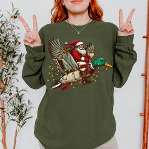Puede incluir: Sudadera verde con una ilustración caprichosa de Papá Noel montado en un pato adornado con luces de hadas. Papá Noel viste su tradicional traje y gorro rojos. El pato tiene plumas verdes y blancas. Un diseño festivo navideño.