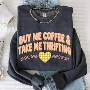 May include: Dark grey sweatshirt with the text "BUY ME COFFEE & TAKE ME THRIFTING" in pink and yellow. The design includes a yellow gingham heart and pink leaf accents. A gold chain necklace and blue jeans are also visible.