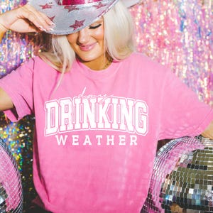 Könnte beinhalten: Pinkes T-Shirt mit dem Text "days drinking weather" in weißen Buchstaben. Das Shirt wird von einer Person mit einem silbernen und rosafarbenen Cowboyhut getragen.