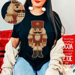 May include: Black t-shirt with a detailed illustration of a Christmas nutcracker soldier. The nutcracker has a tan beard and hair, a large moustache, and is wearing a red uniform with gold accents and a leopard print hat. A smaller inset image of the nutcracker is in the upper left.