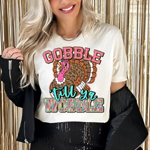 Könnte beinhalten: Weißes T-Shirt mit einem Cartoon-Truthahn-Motiv und dem Text "Gobble 'til ya wobble" in Rosa, Braun und Türkis.