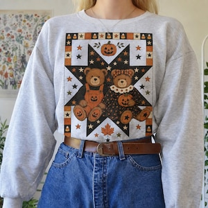 Op de afbeelding: Een lichtgrijs sweatshirt met ronde hals en een grote afbeelding van twee teddyberen in een zwarte ster. De beren dragen herfstkleding en worden omringd door pompoenen, maissnoepjes en esdoornbladeren in een quiltachtig patroon op de borst.