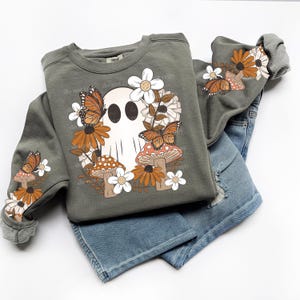May include: An olive green sweatshirt with a ghost graphic surrounded by flowers, butterflies, and mushrooms. The sleeves also feature the same design. Paired with blue denim shorts.