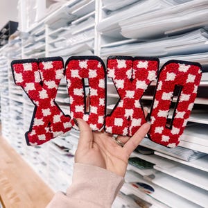 May include: Four decorative letters spelling "XOXO". Each letter is red and white checkered with a black border. The letters are held up in front of a white shelving unit filled with papers.