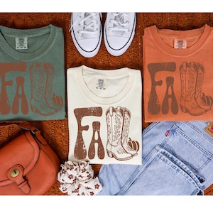 May include: Three t-shirts in sage green, cream, and orange, each with the word "FALL" in brown and a graphic of cowboy boots and a pumpkin. White trainers, a purse, jeans, and a scrunchie complete the autumn-themed ensemble.