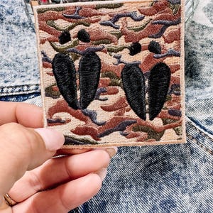 May include: A square embroidered patch featuring a camouflage pattern in shades of tan, brown, and olive green. The patch displays two distinct black embroidered shapes that resemble animal hoof prints centred on the camouflage background.