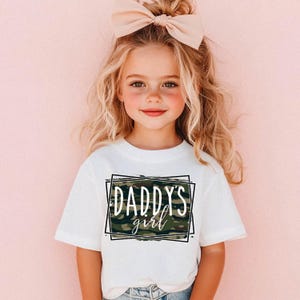 May include: A white t-shirt with the text "DADDY'S girl" in a black frame with a camouflage background. The girl is wearing a large pink bow in her hair and is smiling. The background is a solid pink color.
